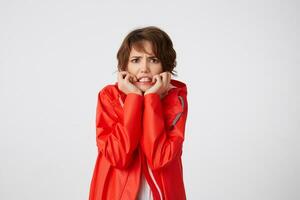 Nice young scared short haired woman in white golf and red rain coat, frowning and fearing looking at the camera, putting hands to cheeks, hears unbelievable news. Standing over white background. photo