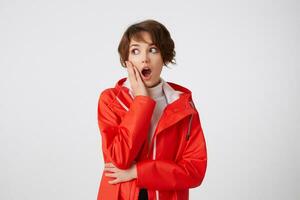 Cute young shocked short haired girl in white golf and red rain coat, looking to the left with wide open mouth, touches the cheek, hears unbelievable gossip. Standing over white background. photo