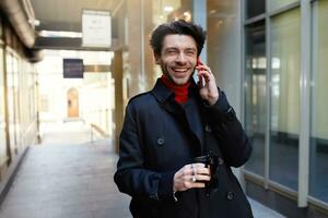 Joyful young attractive brown haired male with trendy haircut laughing happily while having phone conversation with friend, isolated over urban background photo