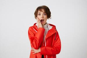 Young cute happy amazed short haired girl in white golf and red rain coat, looking at the camera with wide open mouth, touches the cheek, hears unbelievable news. Standing over white background. photo