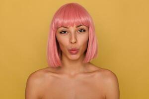 Studio photo of young blue-eyed pink haired woman with natural makeup putting her lips together while looking cheerfully to camera, posing over mustard background