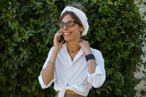 Attractive brunette lady in vintage trendy eyewear and white headband keeping raised hand on her neck and having talk on her smartphone, being in nice mood and smiling happily photo