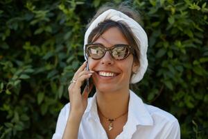 Joyful attractive young lady in vintage eyewear and white headband keeping smartphone in her hand and making call, smiling happily to camera while posing over green garden photo