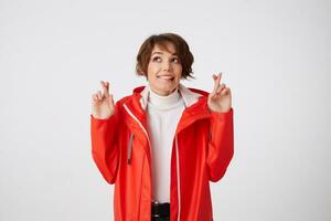 Nice positive young short haired lady in white golf and red rain coat, bites lips, looks up at the copy space, crossed fingers and hopes for luck. Standing over white background. photo