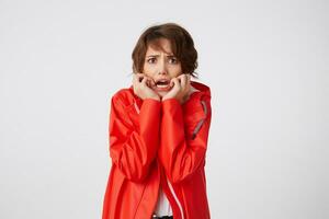 Nice young scared short haired woman in white golf and red rain coat, frowning and fearing looking at the camera, putting hands to cheeks, heard a terrible story. Standing over white background. photo