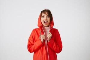 Portrait of young nice happy amazed short haired lady in red rain coat, looking at the camera with wide open mouth, hears cool news. Standing over white background. photo