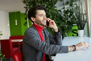 Side view of young handsome unshaved brunette guy dressed in formal clothes sitting at table in cafe and looking at window while having phone conversation photo