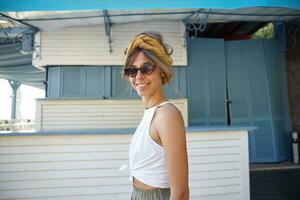 Pretty young brunette woman in vitage sunglasses wearing white top and olive skirt walking over summer terrace while having day off, smiling happily to camera photo