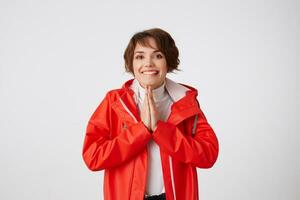 retrato de linda positivo joven corto peludo dama en blanco golf y rojo lluvia abrigo, picaduras labios, mira arriba a el cámara, sonriente, hace oración gesto . en pie terminado blanco antecedentes. foto