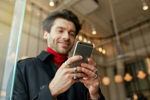 Close-up of raised man's hands with rings keeping mobile phone while posing over city cafe background, texting message to friends and smiling slightly while looking positively on screen photo