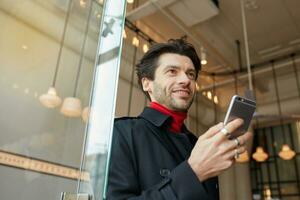 Pleasant looking young attractive dark haired man smiling gladly while keeping smartphone in raised hand, posing over cafe interior in elegant clothes photo