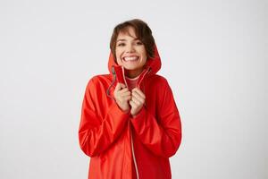 foto de joven bonito alegre corto peludo dama en rojo lluvia abrigo, en general sonriente, disfruta el vida, siente felicidad. en pie terminado blanco antecedentes.