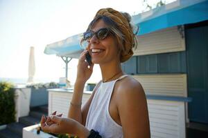 de cerca de encantador joven dama en Clásico de moda Gafas de sol vistiendo casual ropa y venda, hablando en teléfono y sonriente extensamente mientras posando terminado verano café interior foto