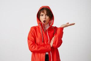 Cute outraged young short haired lady in red rain coat, looks admiringly with wide open mouth in surprice. Standing over white background with crossed arms. photo