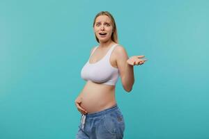 Embarrassed young pretty white-headed woman with natural makeup grimacing her face and raising confusedly palm while posing over blue background, going to be mom soon photo