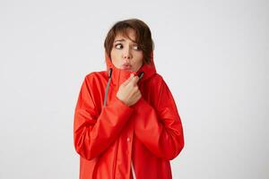 Photo of beauty young frozen and scared short haired woman in red rain coat, frowning and fearing looking up to the left, feels cold, hides in the hood. Standing over white background.