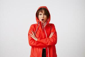 Nice amazed young short haired lady in red rain coat, looks admiringly with wide open mouth. Standing over white background with crossed arms. photo