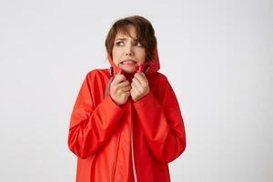 Photo of beauty young scared short haired woman in red rain coat, frowning and fearing looking up to the left, feels cold, hides in the hood. Standing over white background.
