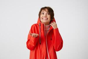 Photo of beauty young smiling short haired woman in red rain coat, looking up to the left, hides in the hood, puts the palm under the rain.Standing over white background.