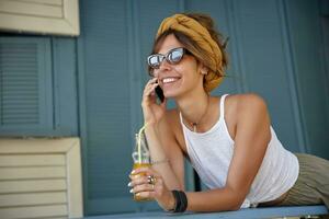 Outdoor photo of young brunette woman in vintage sunglasses wearing casual clothes and mustard headband, posing over summer terrace and drinking lemonade, looking ahead and smiling happily