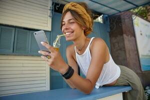 Portrait of attractive young brunette female wearing white top and olive skirt posing over summer terrace, drinking lemonade with straw and typing message to her friend photo