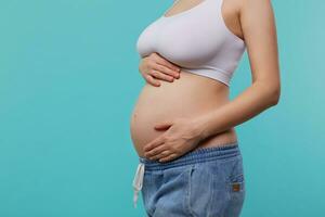 Cropped photo of young pregnant female dressed in white sporty top and jeans holding raised hands on her belly while posing over blue background