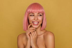 Happy young attractive pink haired female with short haircut touching gently her face with raised hands and smiling pleasantly while looking aside, isolated over mustard background photo