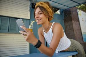 Positive young brunette woman in mustard headband with casual hairstyle drinking orange juice with straw, holding mobile phone in hand and looking at screen while posing over cafe interior photo