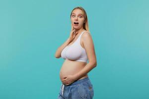 Amazed young blonde long haired pregnant lady looking surprisedly at camera with wide eyes opened and holding hand on her belly, isolated over blue background photo