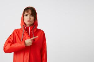 Portrait of young unhappy short haired lady dressed in red rain coat, with a hood on his head, wants to draw your attention, points fingers to the copy space, standing over white background. photo