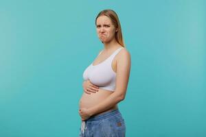 Indoor photo of young white-headed woman with casual hairstyle being pregnant while posing over blue background, twisting her mouth while looking sadly at camera