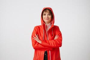 Young smiling cute short haired woman dressed in red rain coat, looking at the camera with a happy expression, standing over white background with crossed arms. Positive emotion concept. photo