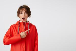 Young nice happy amazed short haired lady in red rain coat, with wide open mouth, hears cool news. Wants to draw your attention, points fingers to the copy space. Standing over white background. photo