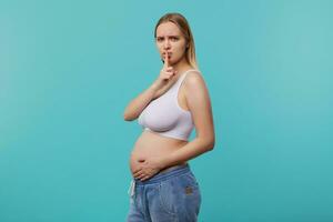 Gloomy young pretty blonde long haired pregnant lady keeping raised index finger on her mouth while looking seriously at camera, isolated over blue background photo