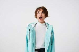 Photo of shocked short-haired curly lady in white golf and light blue rain coat, stands over white background with wide open mouth and surprised expression, looks away with wide open eyes.