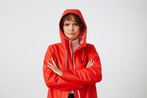 Portrait of young unhappy with rainy weather nice short haired lady dressed in red rain coat, with a hood on his head, looking away with sad expression, standing over white background. photo