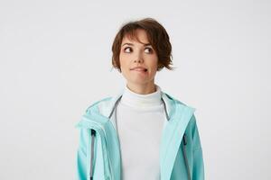 Portrait of young wishful short-haired lady in white golf and light blue rain coat, stands over white background, bites lips and looks away. photo