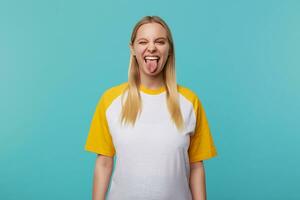 Indoor photo of young pretty long haired blonde female showing happily tongue while making fun, dressed in white and yellow t-shirt while posing over blue background