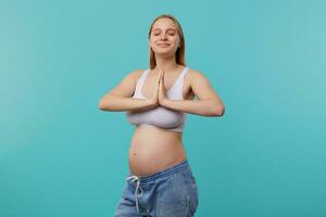 Pleased young attractive blonde pregnant female keeping raised palms together while meditating with closed eyes, being in nice mood while posing over blue background photo