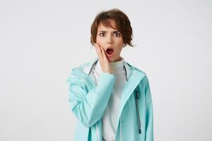 Shocked short-haired young woman in white golf and blue rain coat, stands over white background with wide open mouth and surprised expression, touches cheek, looks at the camera with wide open eyes. photo