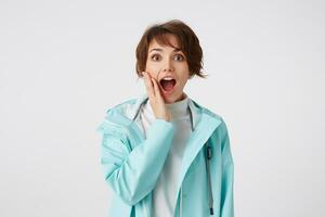 Happy amazed short-haired young woman in white golf and blue rain coat, touches cheek, looks at the camera with wide open mouth and surprised expression, stands over white background. photo