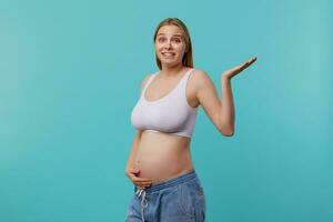 Displeased young pretty white-headed pregnant woman dressed in casual wear raising confusedly palm while looking at camera, standing over blue background photo