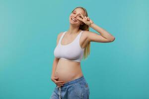 Cheerful young lovely pregnant white-headed female holding her belly with raised hand and showing victory gesture while smiling widely at camera, isolated over blue background photo