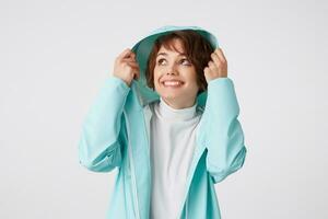 retrato de linda sonriente de pelo corto Rizado dama en blanco golf y ligero azul lluvia abrigo, ocultación debajo el capucha y mirando arriba, soportes terminado blanco antecedentes. foto