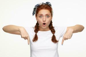 Indoor portrait of young ginger female posing over white background points with a finger to bottom side of the picture photo