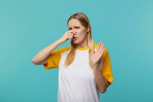 Displeased young attractive long haired blonde female closing her nose with raised hand and frowning face while feeling bad smell, posing over blue background photo