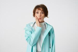 Shocked short-haired young lady in white golf and blue rain coat, stands over white background with surprised expression, touches cheek, looks at the camera with wide open eyes. photo