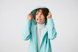 Cute disgusting short-haired curly woman in white golf and light blue rain coat, hiding under the hood from rain and looking up at the left side, stands over white background. photo