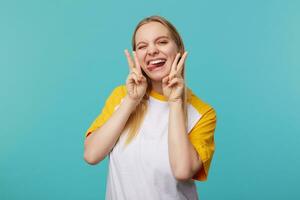 Funny shot of young attractive long haired blonde lady fooling and showing tongue while looking at camera, showing victory sign while standing over blue background photo