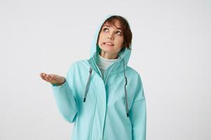 Cute frowning short-haired curly woman in blue rain coat, hiding under the hood from rain and looking up at the rigt side, putting the palm under the rain standing over white background. photo